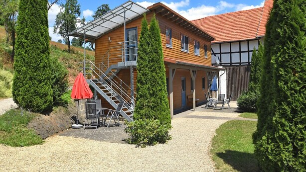 A two-story wooden house with a covered patio, a staircase, and a gravel driveway. - Wohnung in Huddingen mit Terrasse (Bad Wildungen)