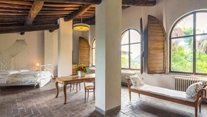 Una habitación rústica con una cama, una mesa de madera con flores, un banco y ventanas arqueadas amplias.