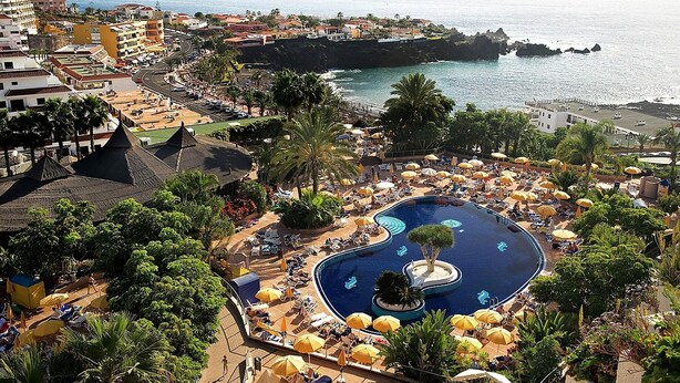 A resort with a large swimming pool surrounded by sun loungers and umbrellas, a view of the sea, and a mix of buildings including a prominent hotel with a red roof.