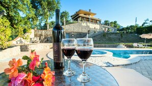 Una mesa con una botella de vino, dos copas de vino tinto y un ramo de flores. Al fondo, una piscina y un edificio con fachada de piedra.