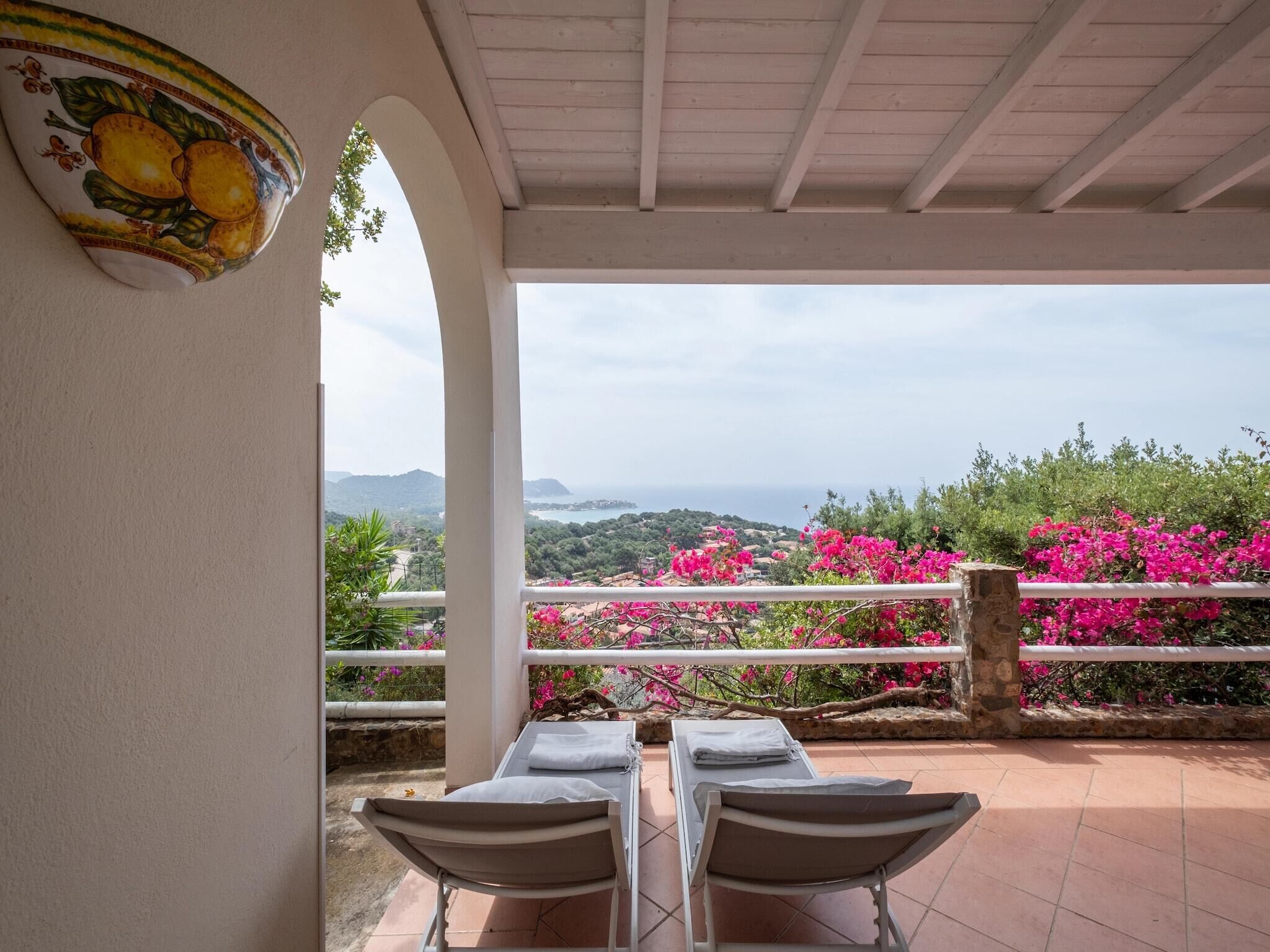 A terrace with two lounge chairs, a decorative wall piece, and a view of the sea and hills.