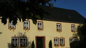 A two-story building with a yellow facade, multiple windows with red frames, and potted plants on the ground floor.
