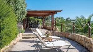A covered outdoor seating area with lounge chairs and a table, surrounded by stone walls and greenery.