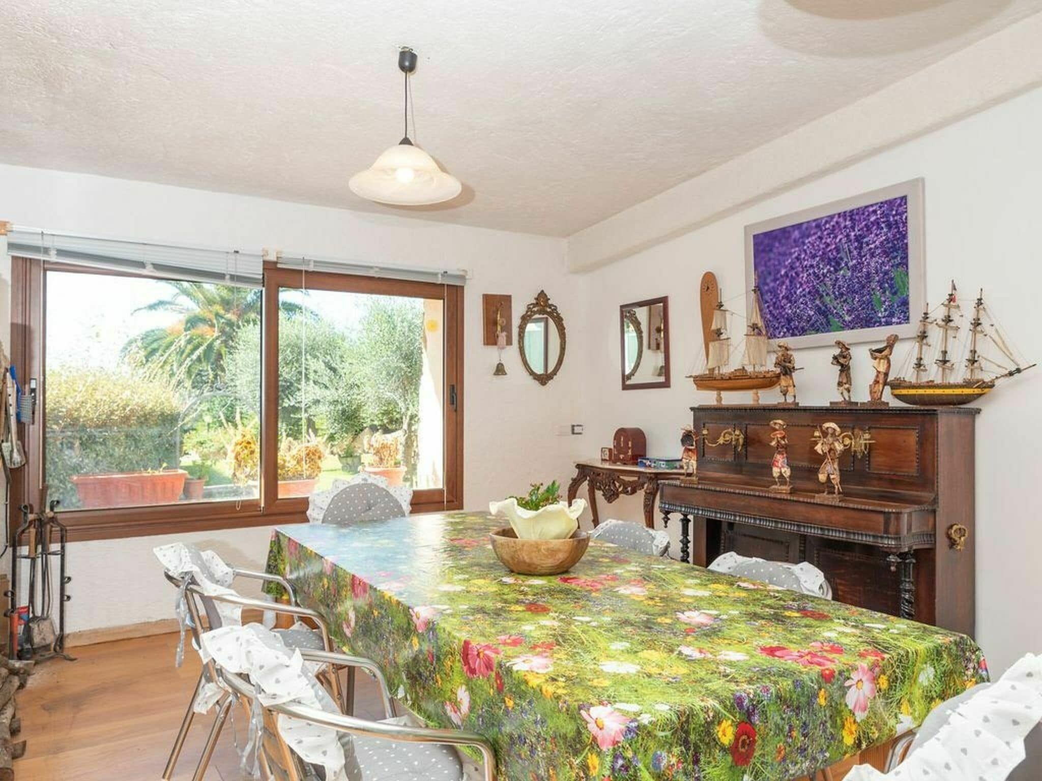 A dining area with a table covered in a floral tablecloth, surrounded by white chairs.