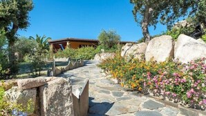 A stone pathway leading to a building with a tiled roof, surrounded by large rocks and flowering plants.