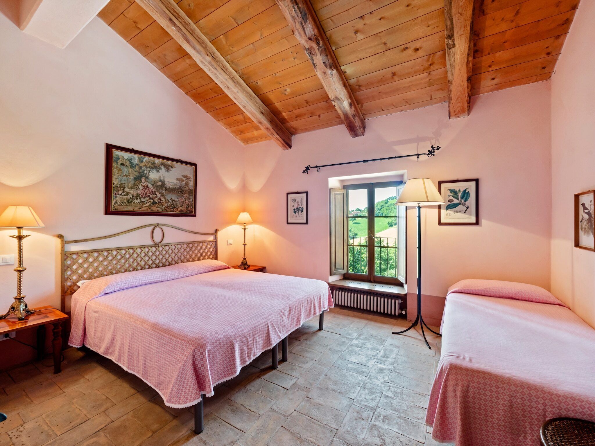 A bedroom with two beds, a wooden ceiling, a window with a view, and a painting on the wall.