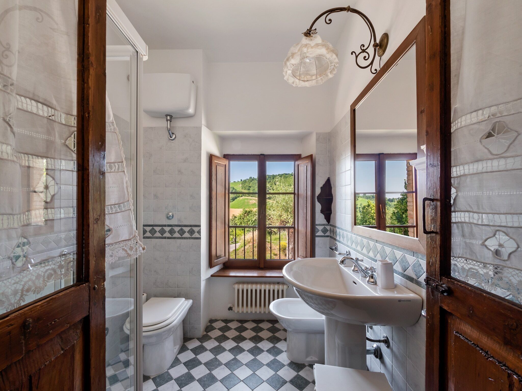 Ein Badezimmer mit Schachbrettmuster-Boden, einem großen Fenster mit Blick auf Grünflächen, einem weißen Waschbecken und einer Toilette.