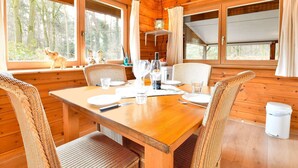 Interieur van een houten hutje met een eettafel voor twee personen, rieten stoelen en uitzicht op het bos door grote ramen.