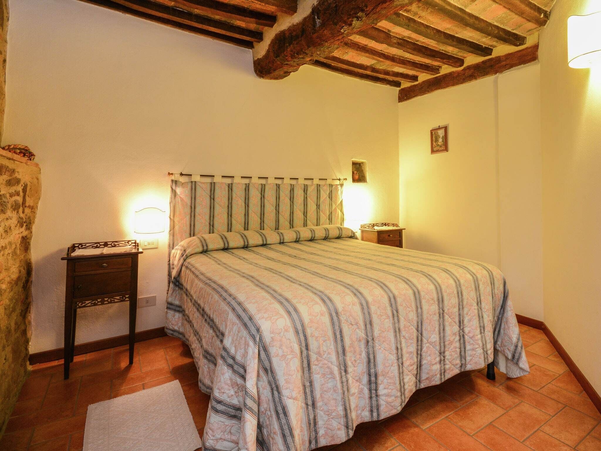 Une chambre à coucher comprenant un lit, deux tables de chevet, une commode, une lampe fixée au mur et un plafond à l’aspect rustique.