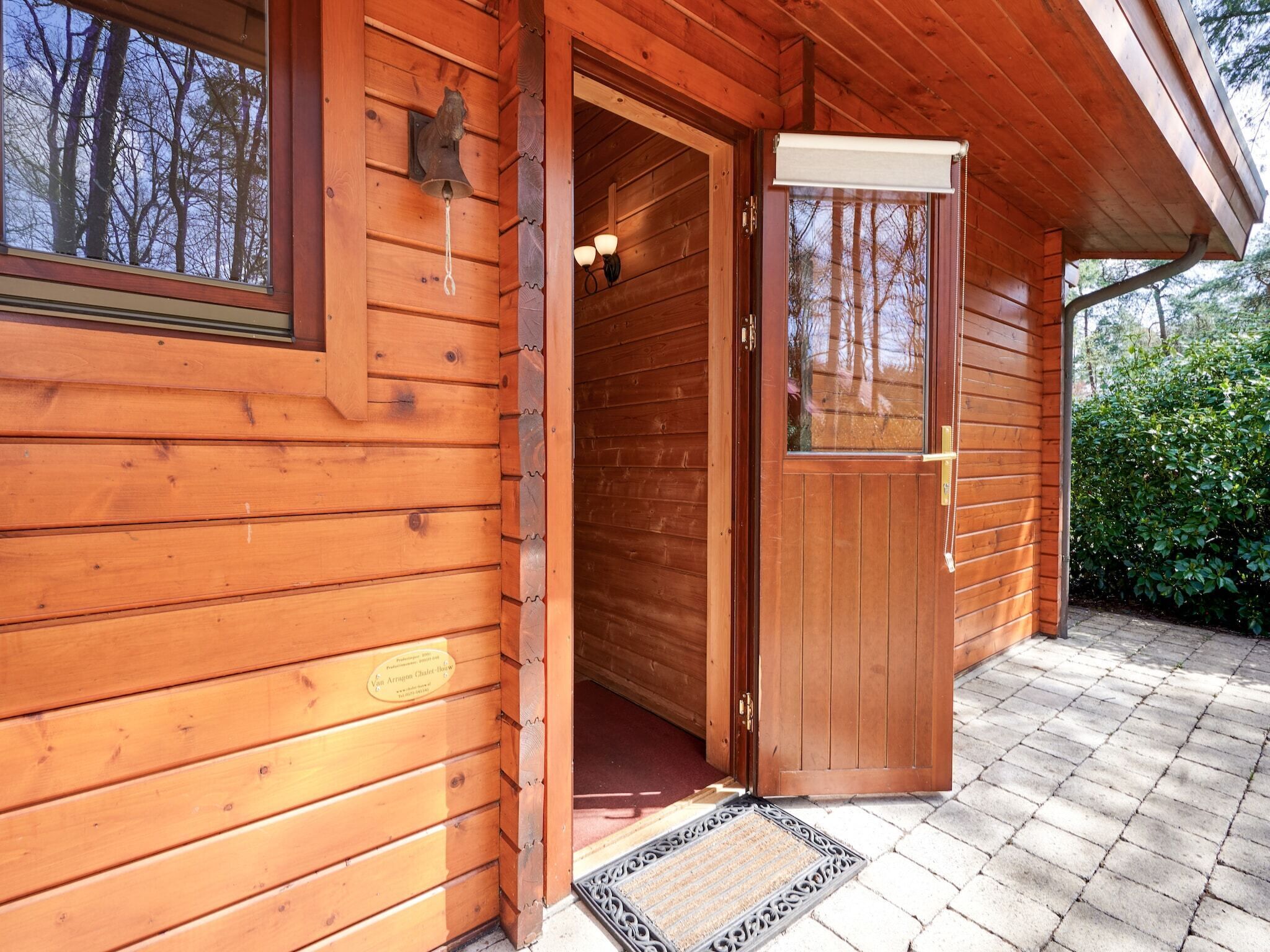 A wooden cabin with a door open, revealing a porch with a doormat and a window overlooking a wooded area.