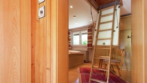 Interior em madeira com escada, cama, cadeira e janela com cortinas.