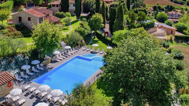 Una gran piscina con tumbonas y sombrillas, rodeada de exuberante vegetación y edificios de piedra.