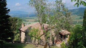 A stone building with a tiled roof, surrounded by lush greenery and a view of distant hills.