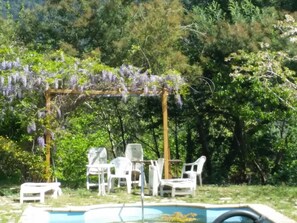 Un área de piscina con una pérgola cubierta de flores moradas, rodeada de sillas y una mesa blancos.