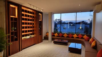 A modern living room with a large window overlooking a body of water, a wooden shelving unit, a patterned floor, and a sectional sofa with colorful cushions.