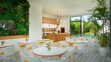 Un espacio interior moderno con una zona de bar, asientos con mesas y sillas de mimbre blanco y un amplio ventanal con vista a una piscina.