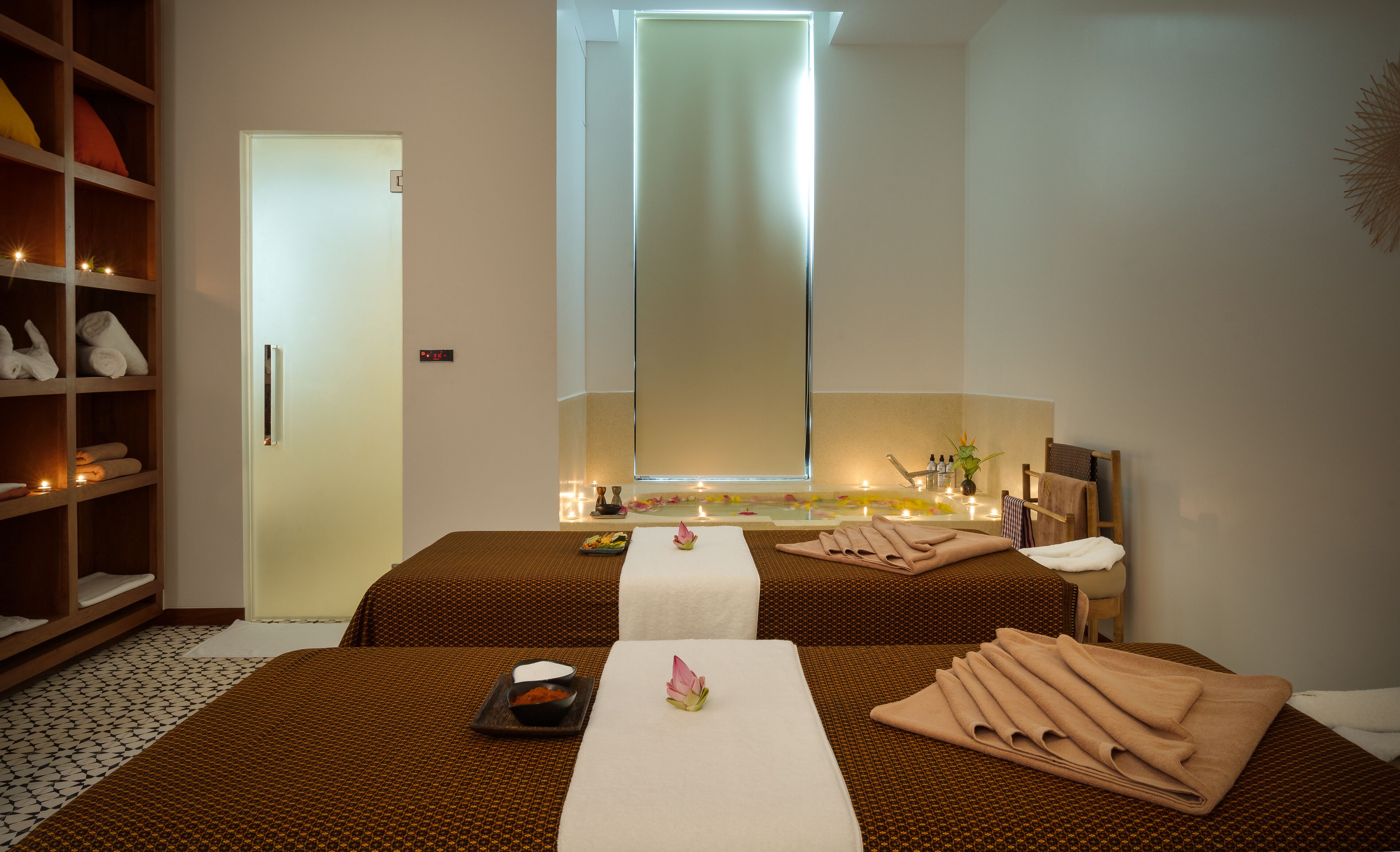 A spa room with two beds, a mirror, a shelf with towels, and a small table with a flower and a bowl.