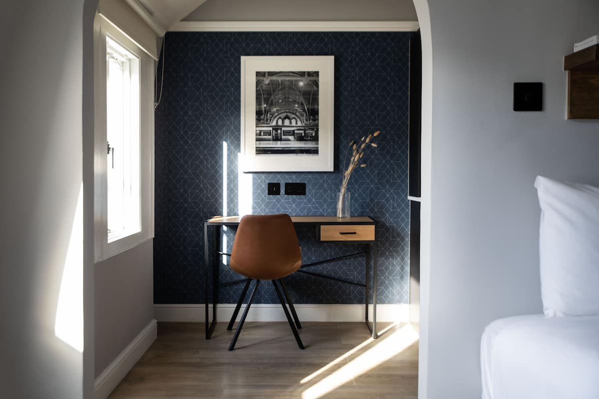 A bedroom with a bed, a desk with a chair, a framed picture on the wall, and a window.