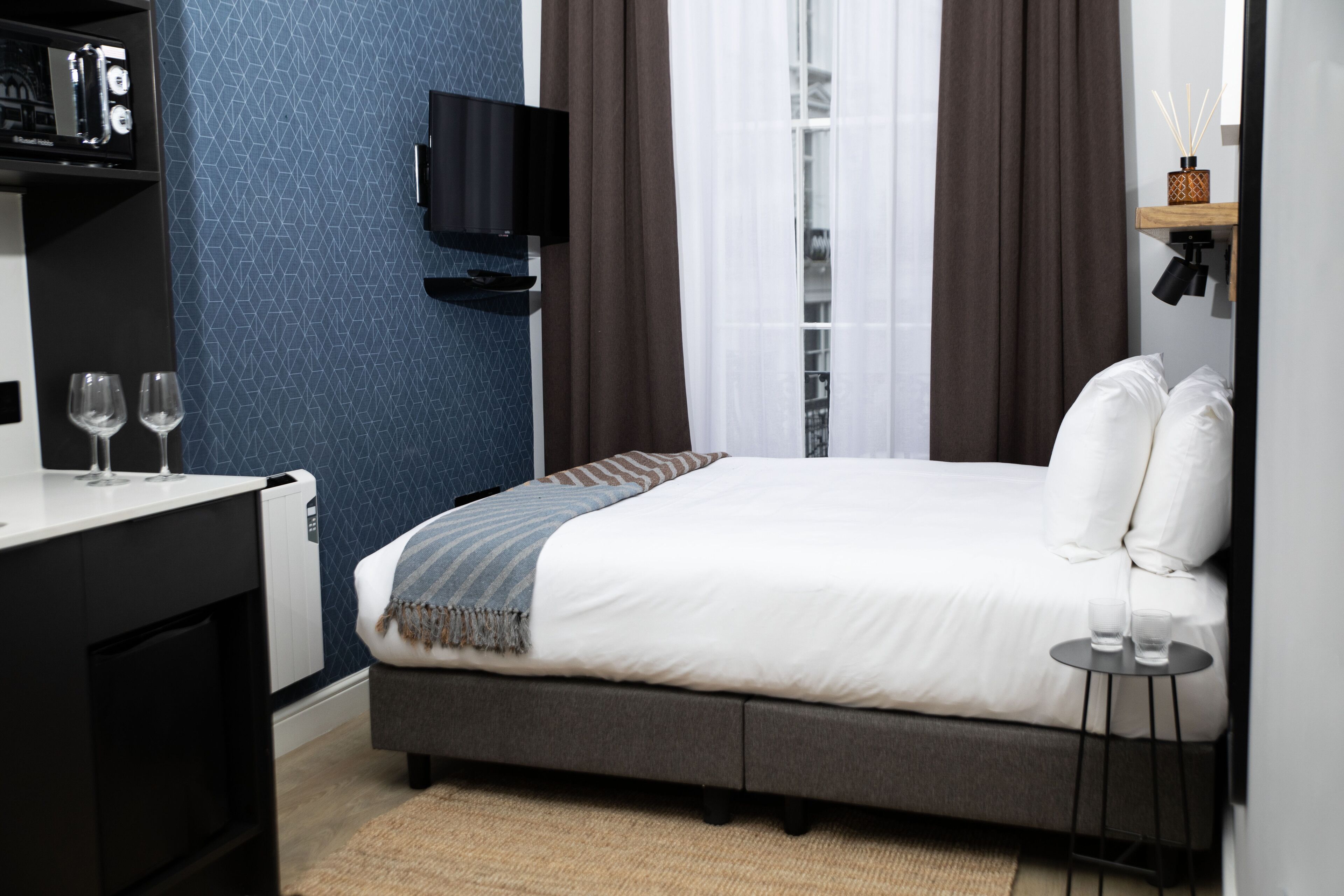 A modern hotel room with a bed, a TV mounted on the wall, a small table with glasses, and a shelf with decorative items.