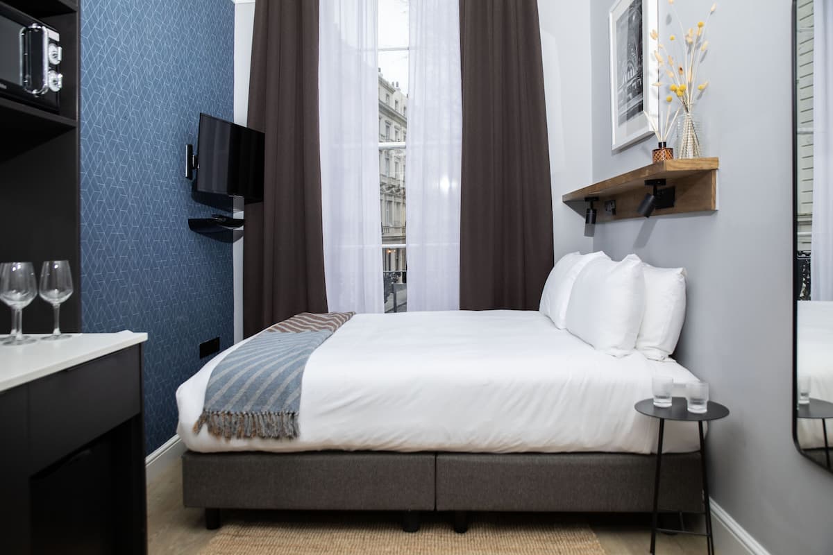 A modern hotel room with a bed, a TV mounted on the wall, a small table with glasses, and a shelf with decorative items.