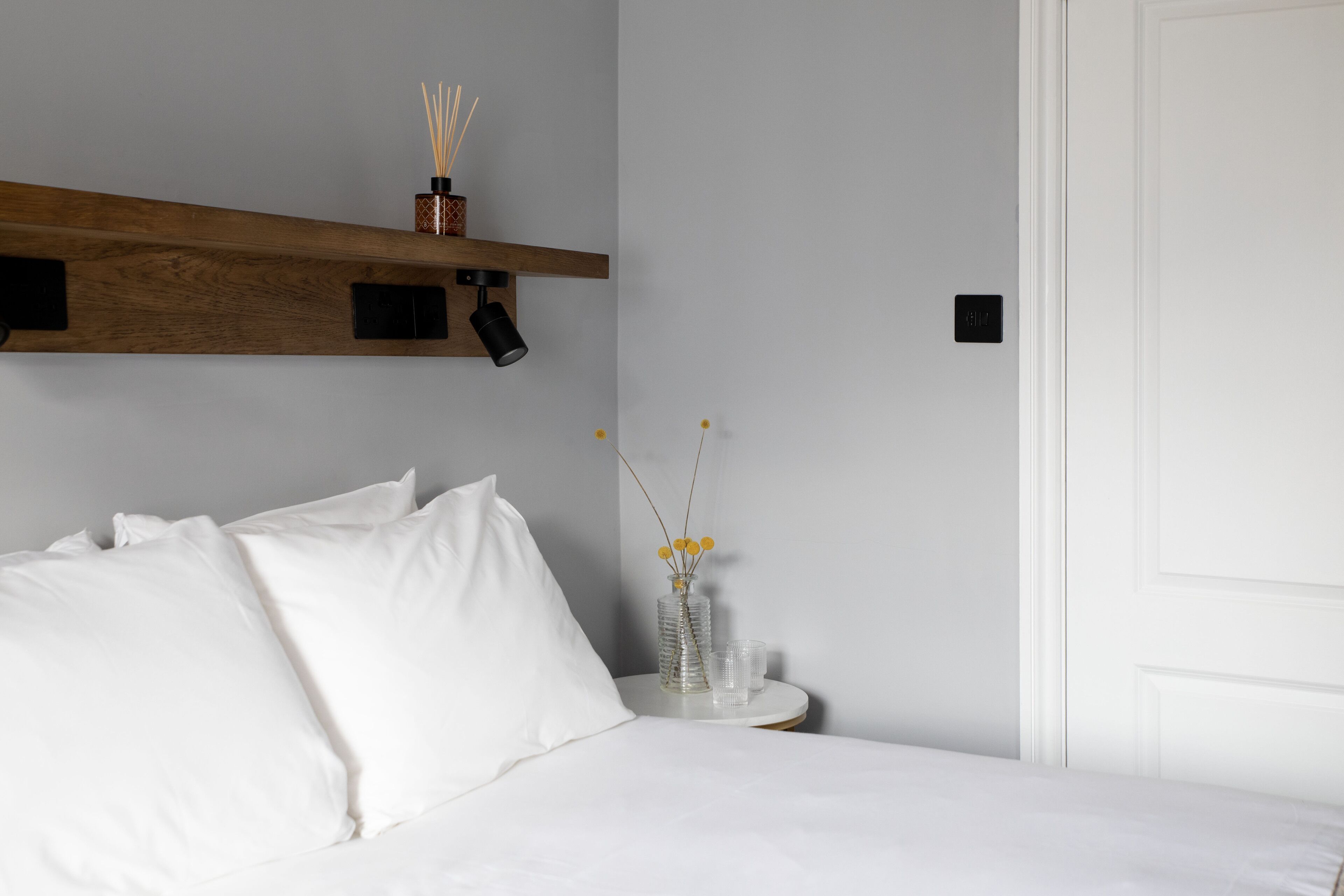 Cama bem arrumada com roupa de cama branca, uma pequena mesa de cabeceira com um vaso transparente e uma vela, e uma prateleira de madeira acima da cama.