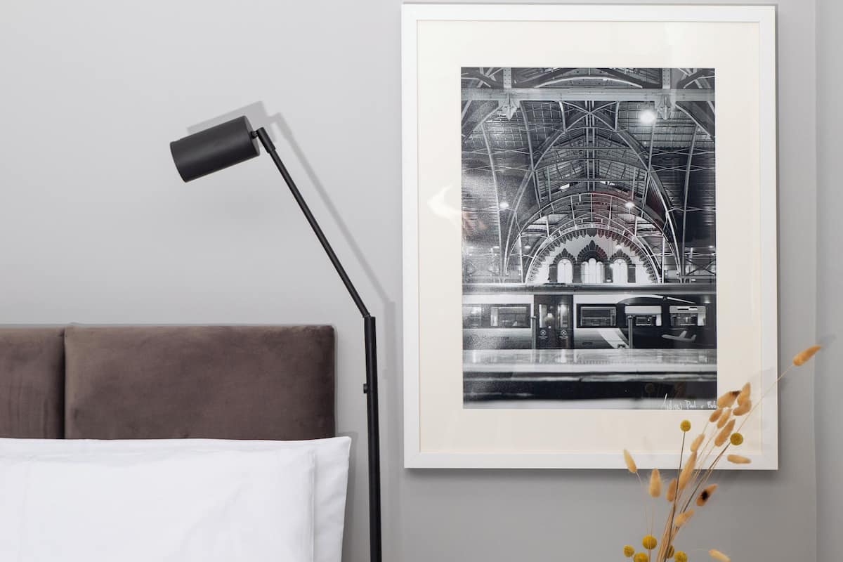 A bedroom with a bed, a bedside table with glasses, a framed black and white photo, and a floor lamp.