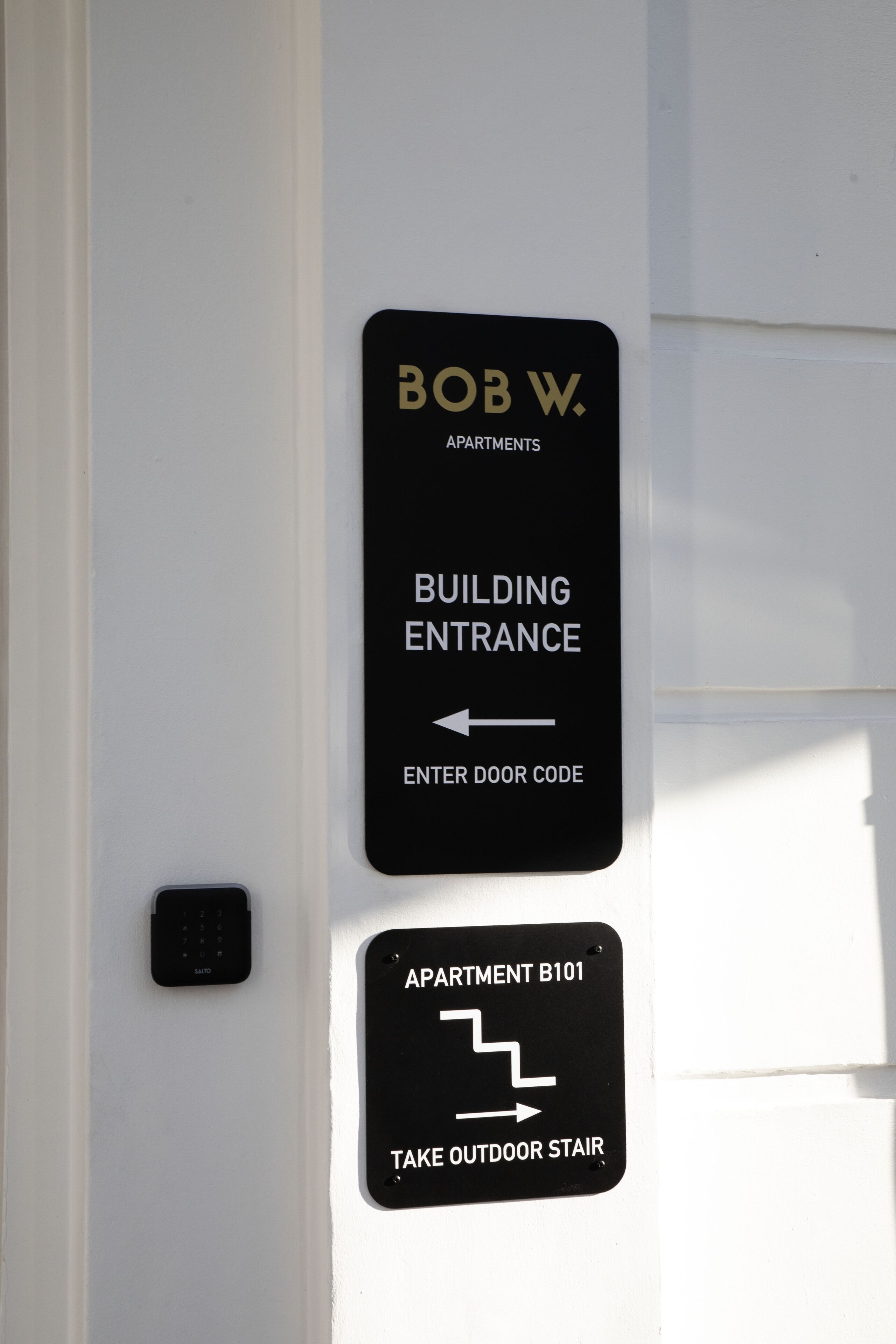 Un panneau d’entrée indiquant l’accès aux appartements Bob W., avec des indications pour trouver l’entrée de l’immeuble et l’appartement B101.