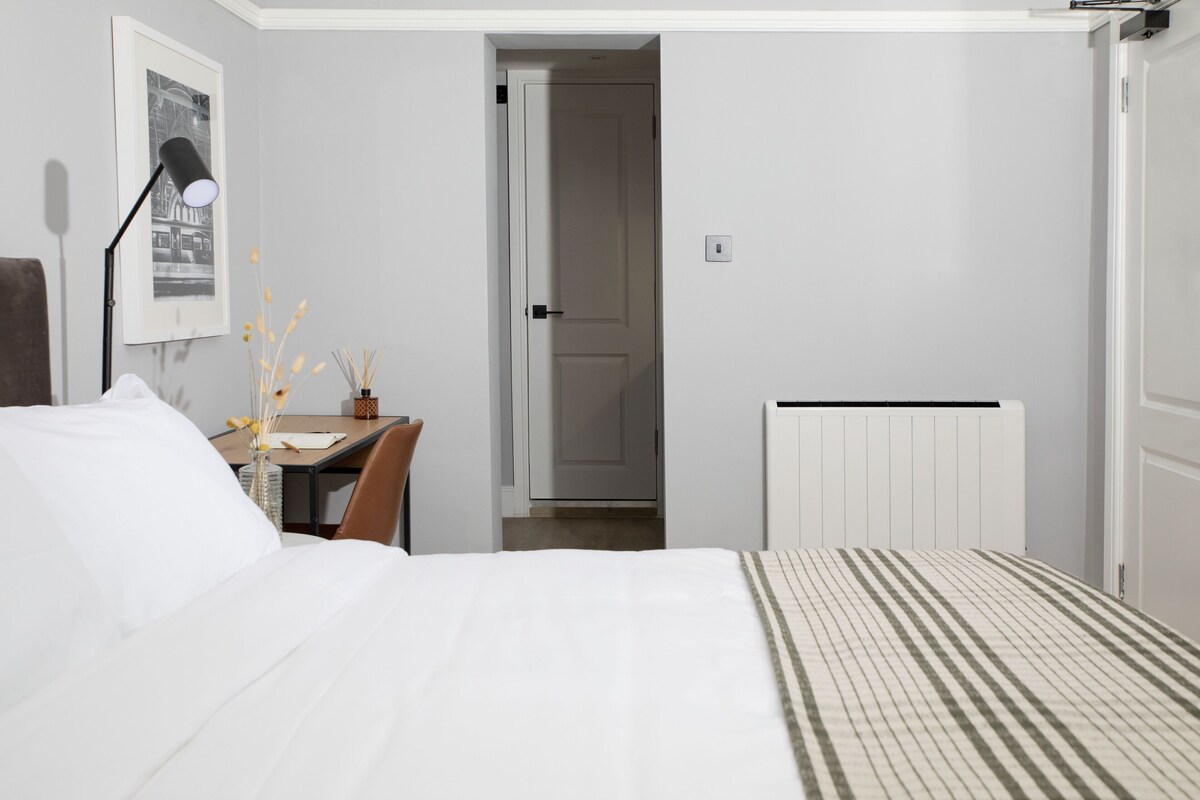 Um quarto com uma cama, uma escrivaninha, uma cadeira, um radiador e uma porta que dá para outro cômodo.