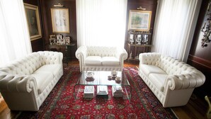 Ein Raum mit drei weißen Ledersofas, die um einen Glastisch angeordnet sind, einem Teppich mit rotem Muster und gerahmten Kunstwerken an den Wänden.