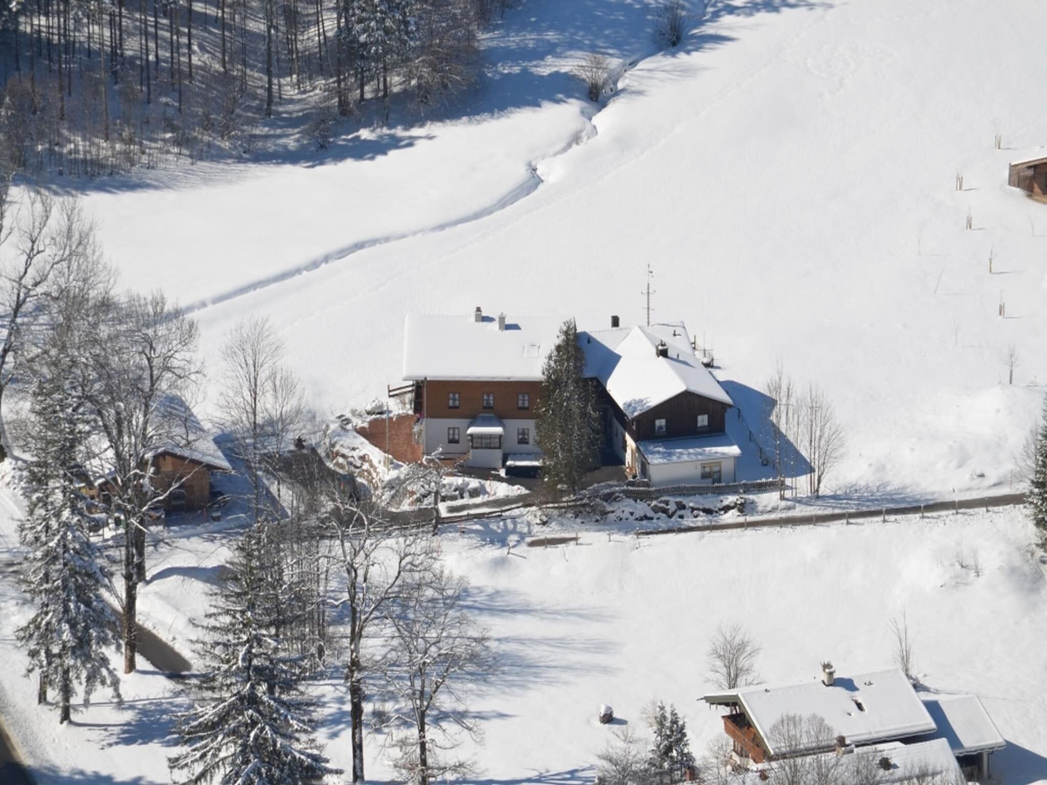 A snow-covered landscape with a large building featuring multiple roofs and a prominent chimney, surrounded by trees and a snow-covered slope.