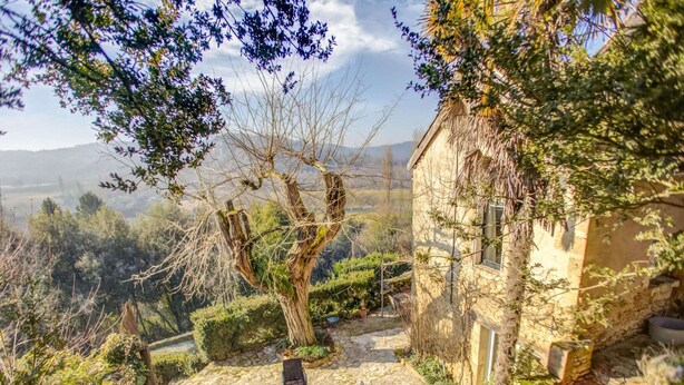 A rustic stone building with a tiled roof, surrounded by a garden with a variety of plants and a tree. There are outdoor seating areas with tables and chairs.