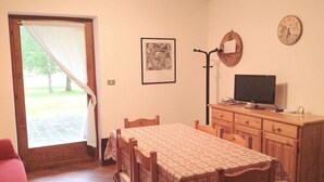 A room with a dining table, chairs, a TV on a wooden cabinet, a framed picture on the wall, and a door leading outside.