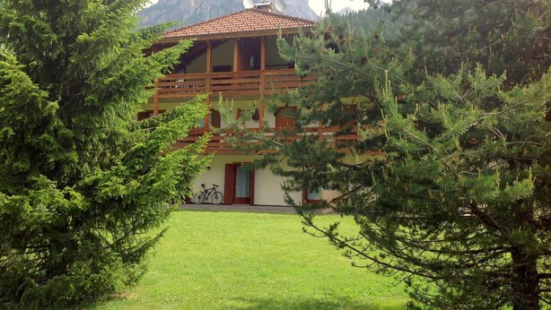 A two-story chalet with a balcony, surrounded by trees and mountains in the background.