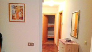 A narrow hallway with a wooden dresser and a mirror on the right, a framed picture on the left, and a door leading to a room with a bed.