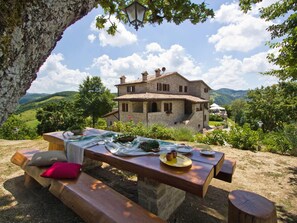 Een stenen huis met een terras, een picknicktafel gedekt met borden en bestek, en een uitzicht op glooiende heuvels.