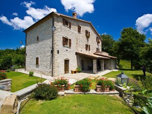 A stone building with a porch, surrounded by a well-maintained garden with red flowers and a clear blue sky. - Belvilla by OYO La Rocca (Apecchio)