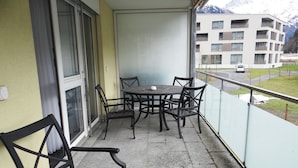 A balcony with a table and chairs, a window, and a view of a building and mountains.