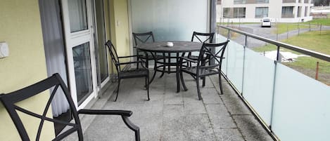 Een balkon met een tafel, stoelen, een raam en uitzicht op een gebouw en bergen.