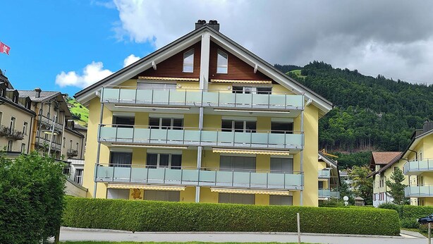 Ein mehrgeschossiges Gebäude mit Balkonen vor einer grünen Hügellandschaft und blauem Himmel.