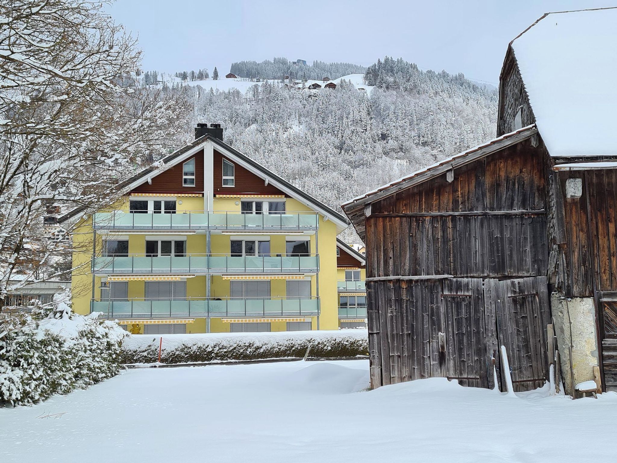 A multi-story yellow building with balconies, a wooden barn, and a snowy landscape with a hill in the background.