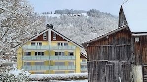A multi-story yellow building with balconies, a wooden barn, and a snowy landscape with a hill in the background.