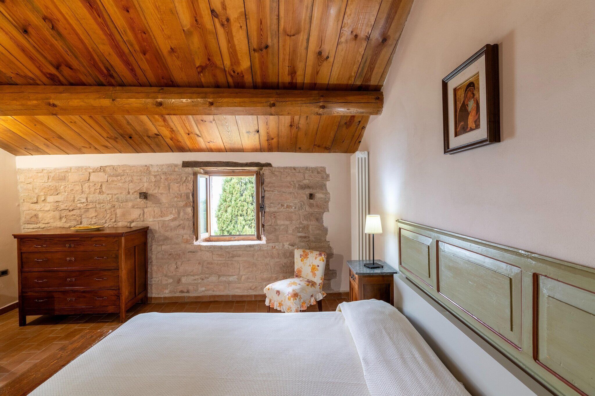Une chambre avec un plafond en bois, un mur en pierre, une fenêtre, un lit, une chaise, une commode et une lampe.