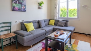 A modern living room with a grey sectional sofa, a small table, and a chair. There is a window with a view of greenery outside. - Pretty holiday home with garden and hot tub (Bédoin)