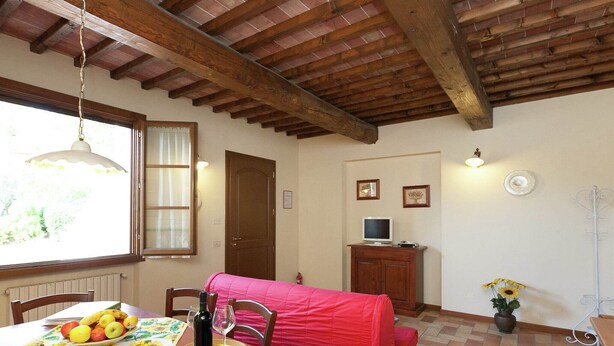 A cozy room with a wooden ceiling, a dining table with a fruit bowl, a wine bottle, and a red chair. There's a window with curtains, a door, and a small cabinet with a TV. - Belvilla by OYO Giaggiolo (Stabbia)