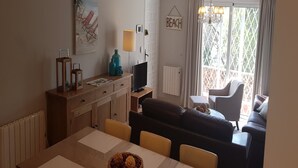 Una sala de estar acogedora con sofá, mesa de comedor y televisor. Se observa un cartel con la palabra 'BEACH' en la pared y una ventana con cortinas.