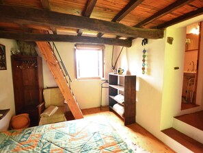 Una stanza con soffitto spiovente, un letto con copriletto, una finestra, uno scaffale con teiera e una scala che conduce al bagno.