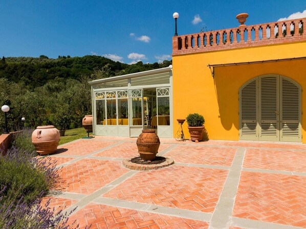 Eine Terrasse mit Terrakottafliesen, ein Wintergarten mit Glastüren und ein gelbes Gebäude mit Ziegeldach.