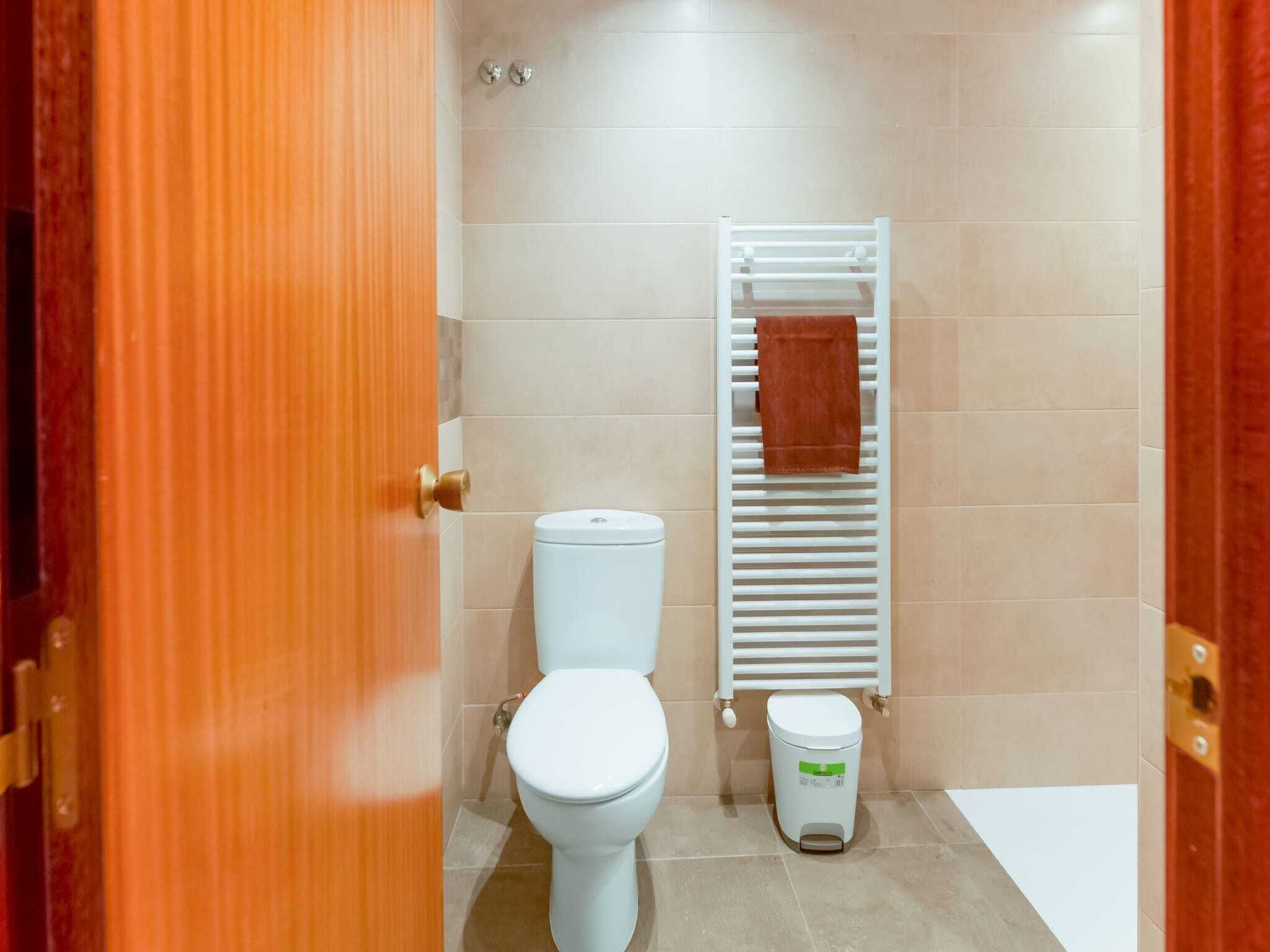 Un baño con inodoro, un perchero blanco con toalla y un cubo de basura.