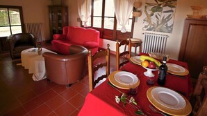 A cozy living room with a red sofa, a brown leather ottoman, and a dining table set for two with plates, glasses, and a bottle. - Quaint cottage with pool (Anghiari)