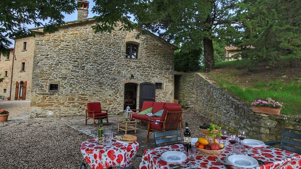 An outdoor dining area with stone walls, red and white floral tablecloths, wicker chairs, and a stone building in the background. - Quaint cottage with pool (Anghiari)